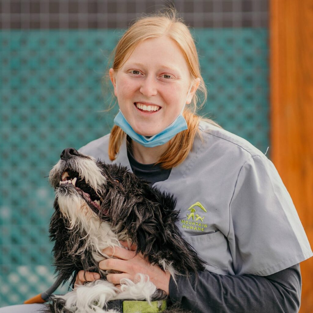 Bailey Gould in her Mountain Humane uniform smiling at the camera with a scruffy dog in her lap looking up at her